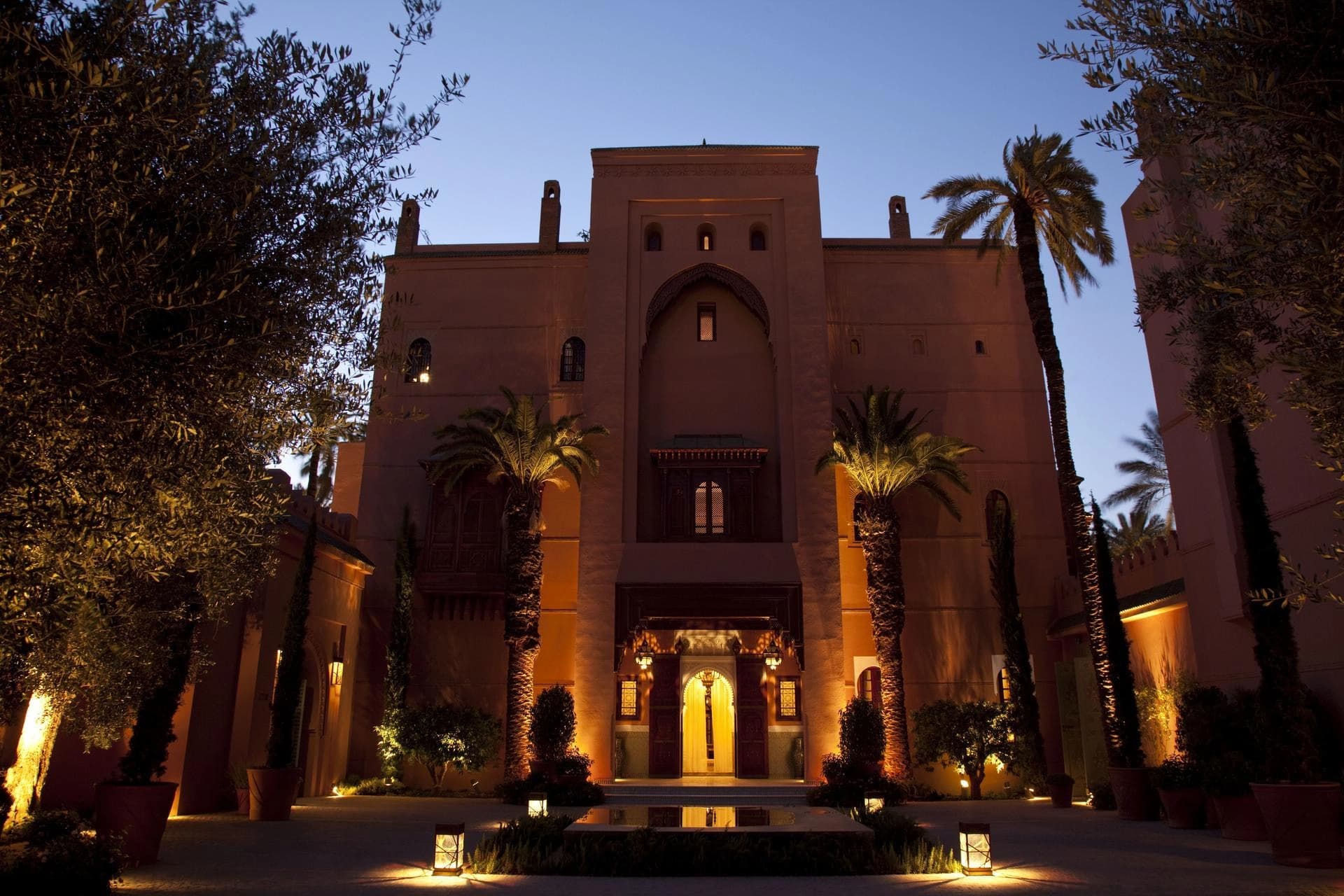 Façade d'un hôtel de style marocain en terre cuite, richement illuminée de lumières chaudes au crépuscule. Des palmiers encadrent l'entrée voûtée, et une fontaine réfléchit la lumière dans la cour sous un ciel bleu foncé.