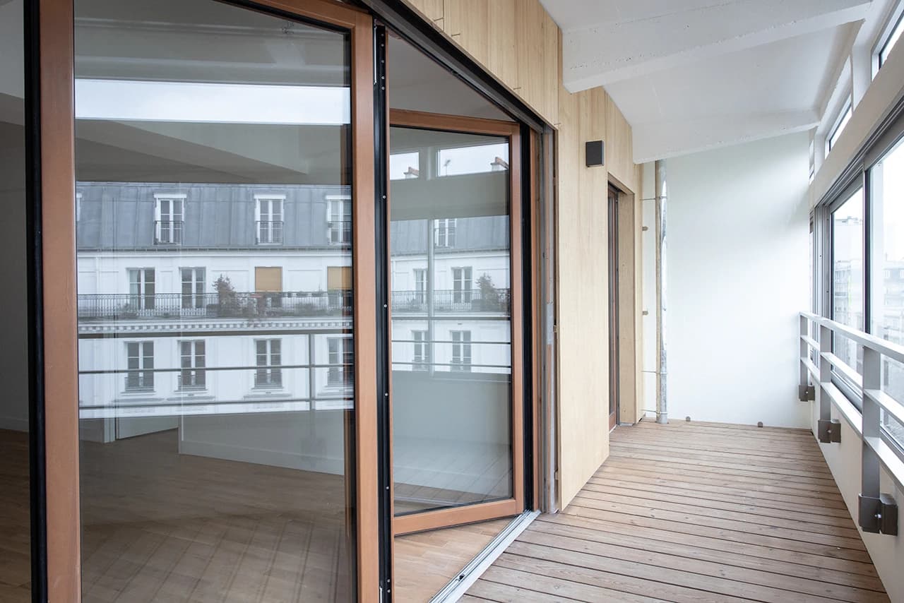 Balcon moderne avec plancher en bois et balustrade grise, ouvert via de grandes baies vitrées coulissantes à cadre bois, reflétant un immeuble haussmannien blanc et l'intérieur d'une pièce vide.