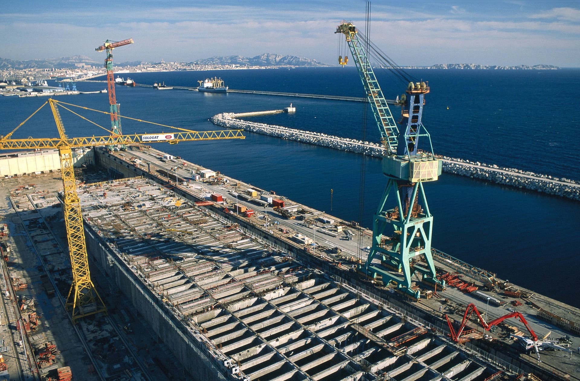 Vue aérienne d'une immense cale sèche en béton en construction dans un chantier naval, flanquée de grues industrielles jaunes et bleu-vert. La mer bleue avec des brise-lames et une ville côtière lointaine forment l'arrière-plan.
