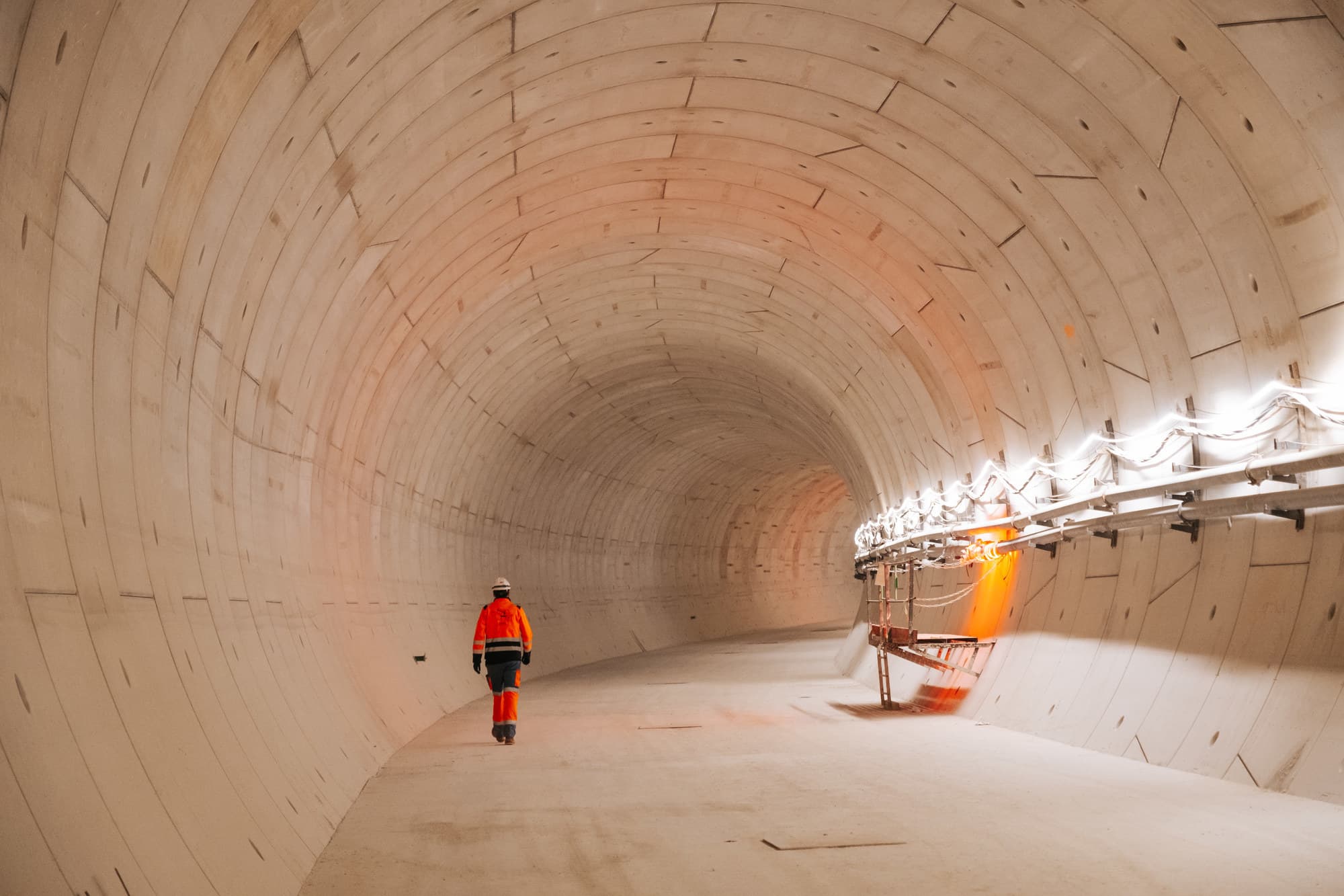Un travailleur en tenue de sécurité orange et casque blanc marche dans un long tunnel en béton clair, fraîchement construit. Des lumières blanches et un halo orange vif éclairent l'intérieur voûté.