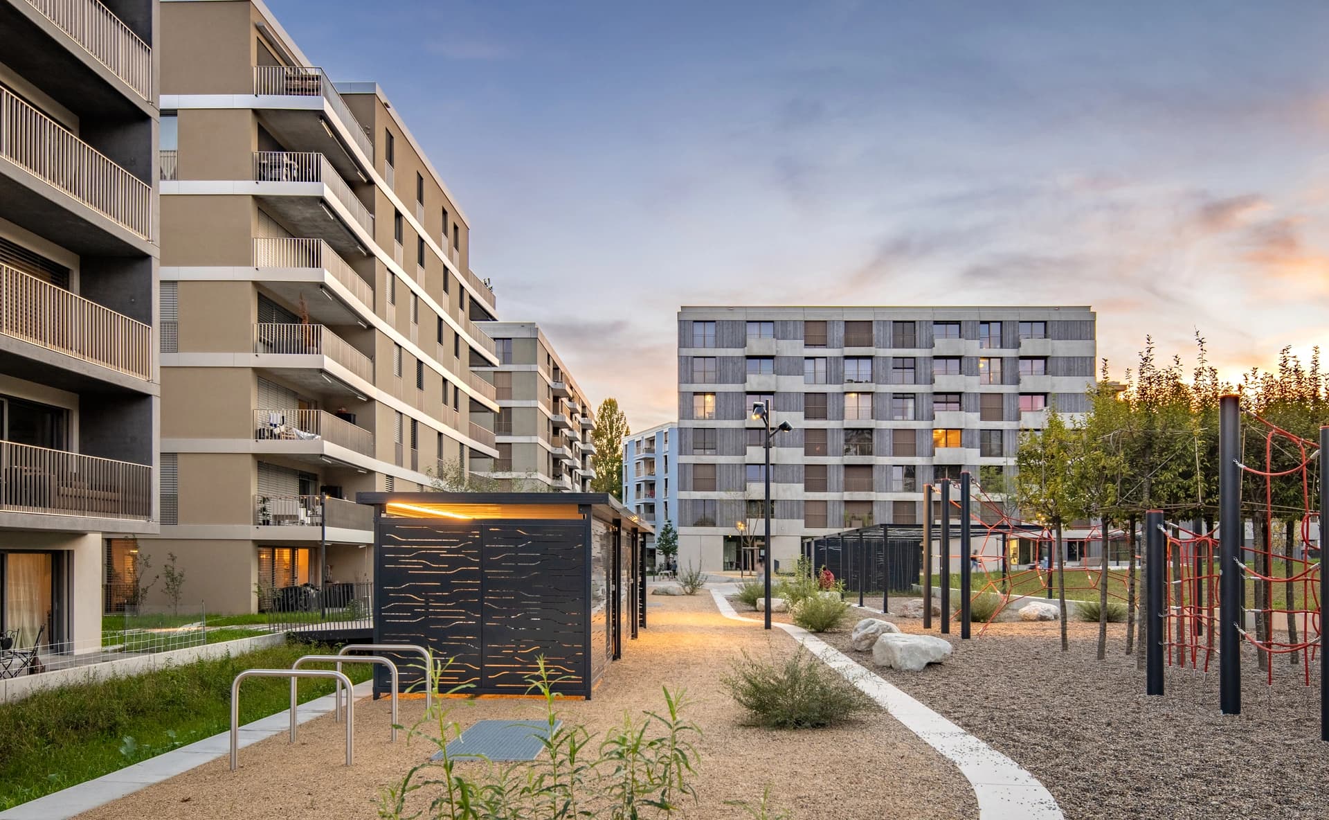Vue d'un complexe résidentiel moderne avec des immeubles aux façades beiges et grises, un chemin de gravier, un espace vert, et une aire de jeux avec des cordes rouges au premier plan, sous un ciel de crépuscule aux couleurs bleu-orange.