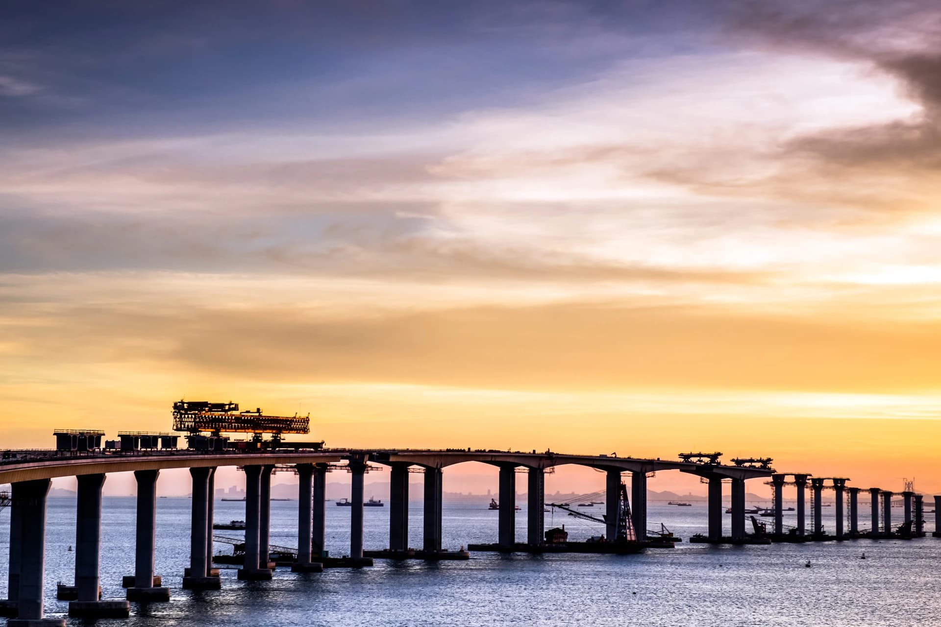 Pont long en construction, silhouetté contre un ciel de coucher de soleil dramatique allant du bleu au violet et au jaune-orange. Des équipements de construction sont visibles sur le pont et dans l'eau bleue en contrebas, indiquant des travaux en cours.