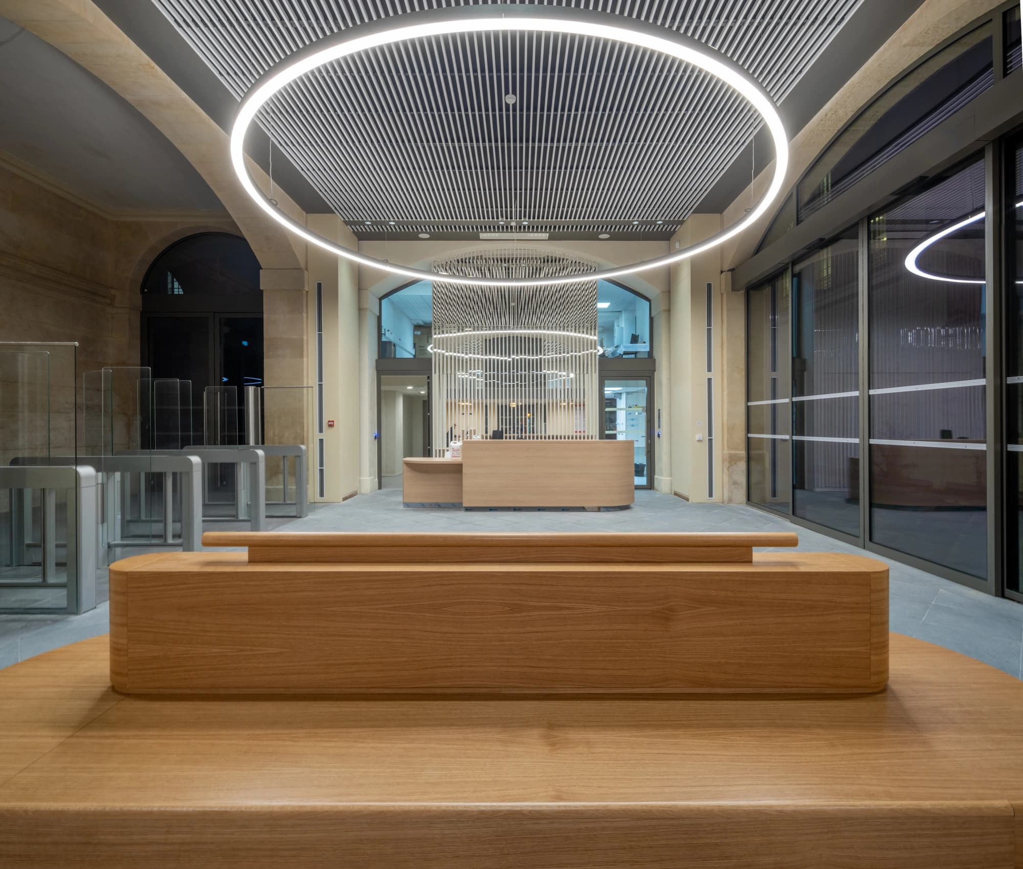 Intérieur moderne et lumineux d'un hall d'accueil présentant un grand banc en bois clair au premier plan, face à des tourniquets de sécurité et un comptoir de réception. L'espace est éclairé par des luminaires LED circulaires sous un plafond à lattes.