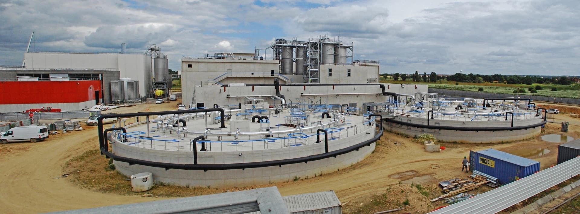 Vue aérienne d'une station d'épuration ou usine de traitement moderne en construction, avec de grands bassins circulaires en béton, des bâtiments industriels gris et rouges, des tuyaux complexes, sur un sol terreux sous un ciel nuageux.