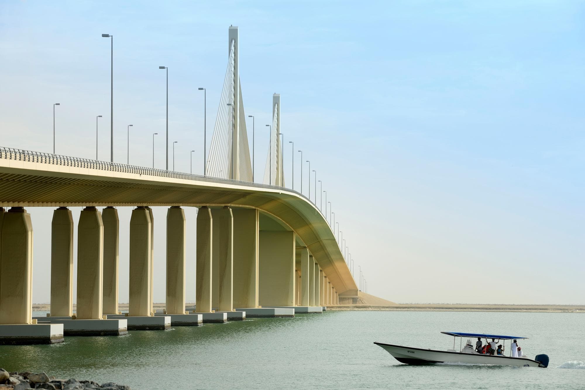 Un grand pont haubané beige clair avec de hauts pylônes et des lampadaires s'étend en courbe au-dessus d'une étendue d'eau vert-bleu sous un ciel pâle. Un petit bateau à moteur blanc avec un auvent bleu et plusieurs passagers navigue sur l'eau au premier plan.