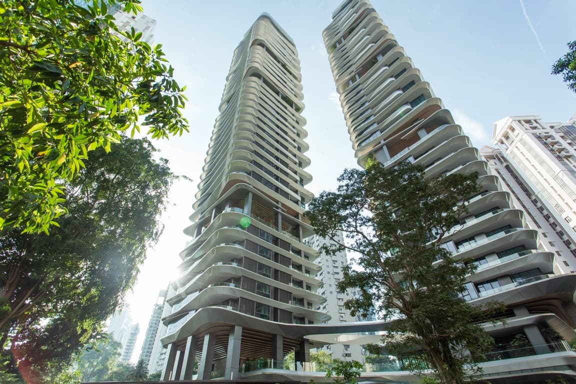 Deux tours modernes aux façades blanches ondulantes et balcons incurvés s'élèvent sous un ciel bleu clair, entourées d'arbres verts luxuriants.