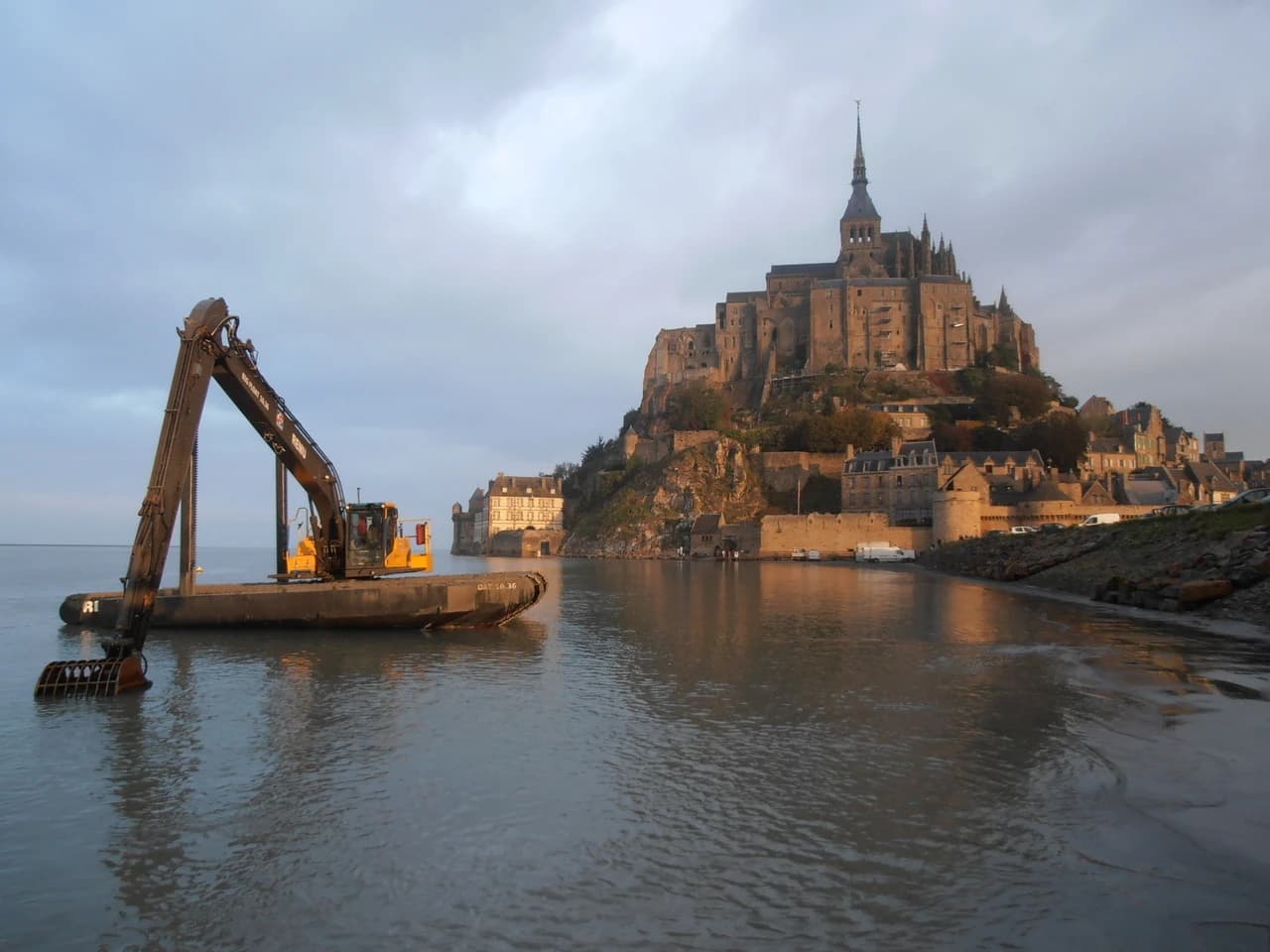 Un excavateur amphibie jaune et noir flotte au premier plan sur l'eau sombre, tandis que l'abbaye et le village du Mont Saint-Michel, majestueux et baignés de lumière dorée, s'élèvent en arrière-plan sous un ciel nuageux.