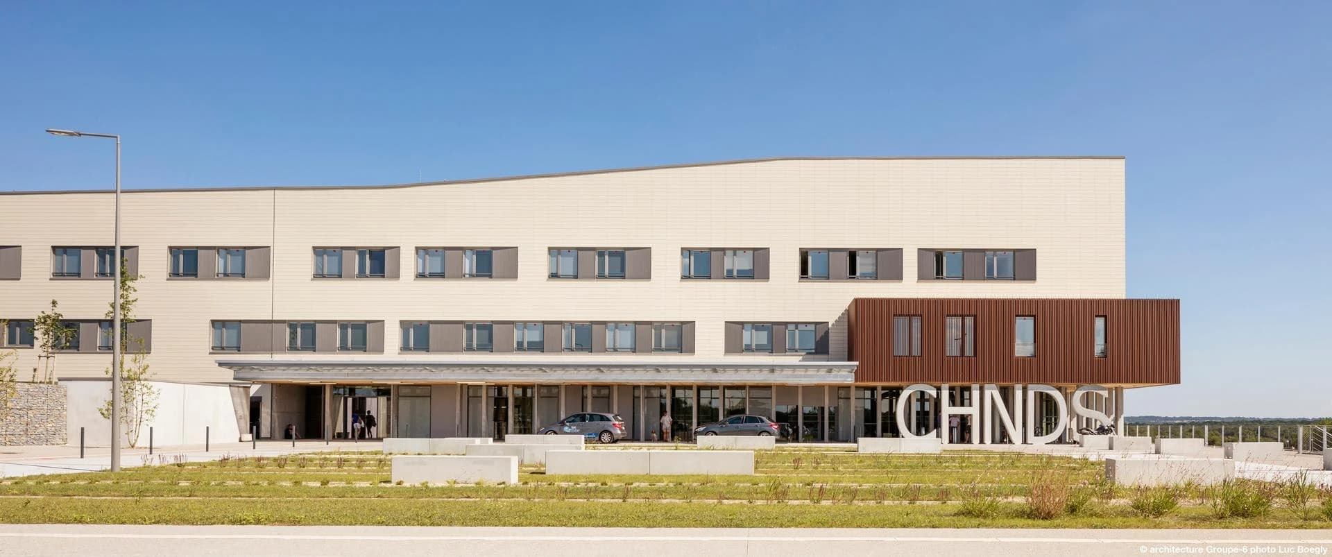 Grand bâtiment moderne avec des façades principales beige clair et une section marron foncé, présentant des rangées de fenêtres avec des volets gris sous un ciel bleu éclatant. Les lettres blanches géantes "CHINDSE" sont visibles devant l'entrée du bâtiment, sur une pelouse avec des blocs de béton.