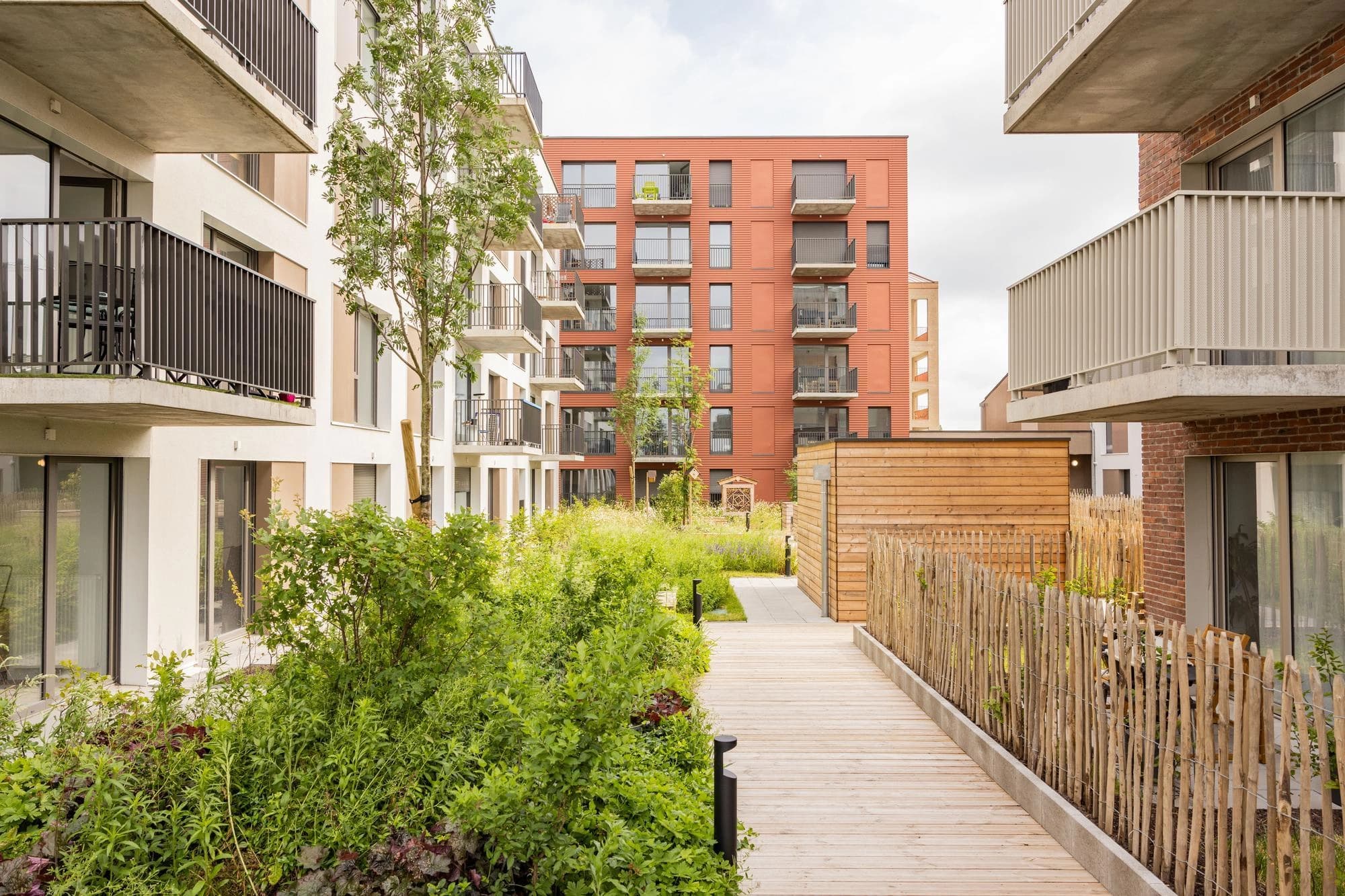 Un espace vert aménagé au cœur d'un complexe résidentiel moderne, avec une allée en bois clair traversant une végétation luxuriante. Des immeubles aux façades blanches et terre cuite, agrémentées de nombreux balcons, entourent ce jardin urbain sous un ciel lumineux.