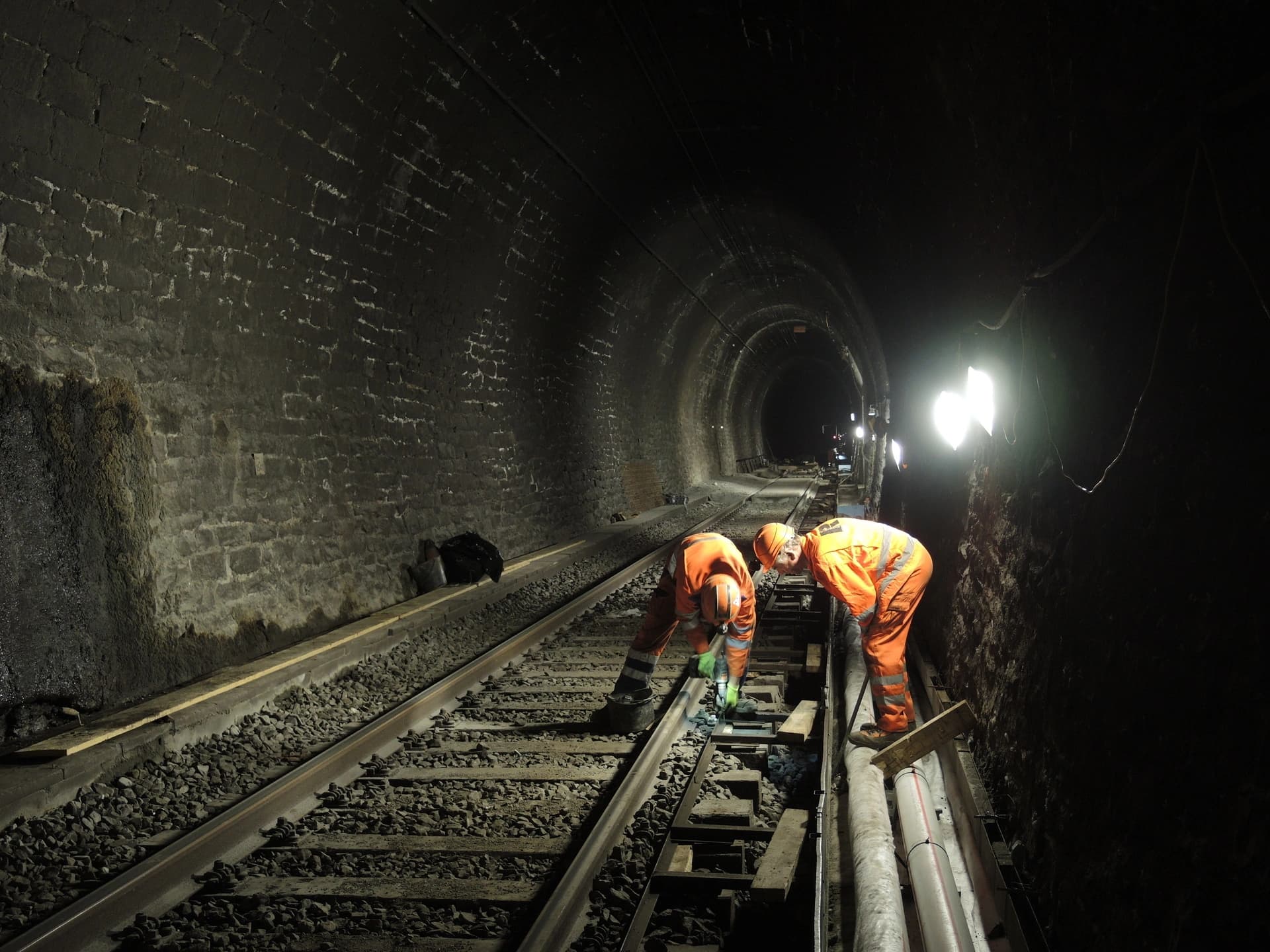 Deux ouvriers en tenues de sécurité orange travaillent sur des voies ferrées à l'intérieur d'un tunnel sombre aux murs de briques, éclairés par de puissantes lampes de travail.