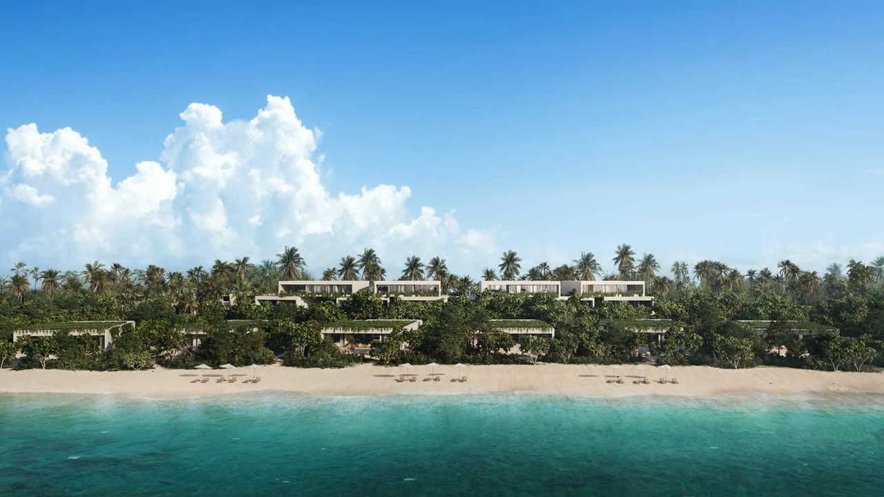 Aerial view of modern villas nestled among palm trees on a tropical white sand beach with turquoise water under a blue cloudy sky.